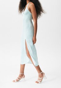 Vaaleansininen midi-mekko, jossa on kukkakuvio, halterneck ja sivuhalkio; yhdistetty valkoisiin remmikkäisiin, paksukorkoisiin sandaaleihin.