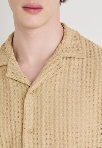 Beige gebreide shirt met een kraaghalslijn, een gestructureerd patroon, natuurlijke vezels en knoopdetails aan de voorkant.
