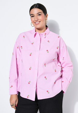 Lächelnde Frau mit dunklen Haaren trägt ein rosa gestreiftes Hemd mit kleinen roten Blumenmustern und schwarze Hosen vor weißem Hintergrund.