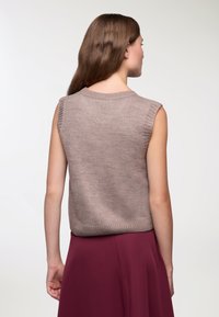 Bezramionowy, taupe'owy sweter dzianinowy z prążkowanym dekoltem i otworami na rękawy, w połączeniu z bordową spódnicą. Materiał wydaje się gładki i teksturowany.