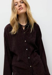 Blonde Frau, die ein lockeres, dunkelbraunes Kordhemd mit Knöpfen und eine schwarze Perlenkette vor einem schlichten, hellen Hintergrund trägt.