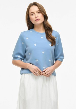 Donna che indossa una maglietta blu a maniche corte con motivi a cuori bianchi e una gonna bianca, in piedi con le mani leggermente intrecciate.