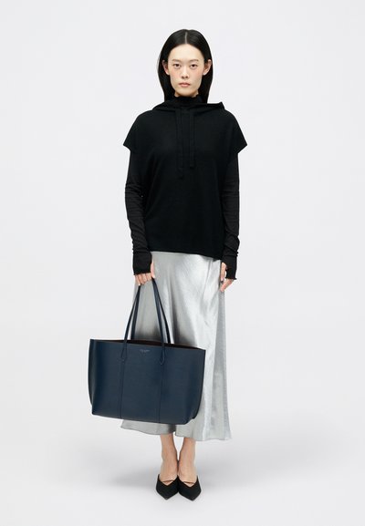 Hoodie preto de manga curta, saia metálica prateada, sapatos planos pretos de bico; bolsa grande de couro azul-escuro com duas alças.