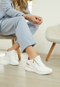 Baskets blanches avec un design texturé, des accents dorés et un talon rouge. Dotées d'une fermeture éclair sur le côté et d'une semelle compensée pour plus de hauteur.