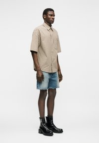 Hombre con camisa de manga corta beige a cuadros, pantalones cortos de mezclilla azul claro con bordes desgastados y botines de charol negro, de pie de perfil.