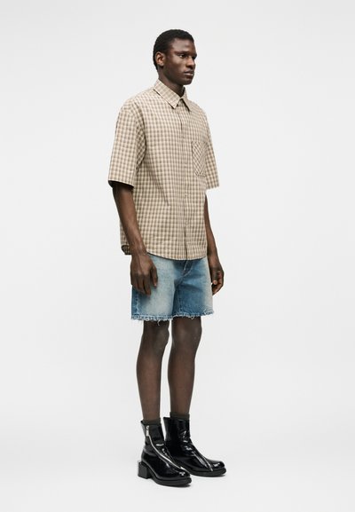 Homme portant une chemise à manches courtes beige à carreaux, un short en jean bleu clair aux bords effilochés, et des bottines noires en cuir verni, debout de profil.