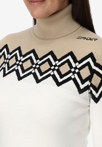 Beige coltrui met een geometrisch zwart-wit patroon over de borst. Gemaakt van een zachte breimateriaal met een geribde kraag.