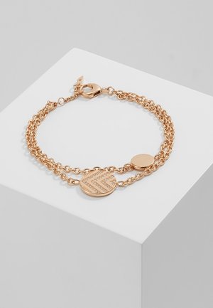 Bracelet à double chaîne en or rose avec un pendentif rond présentant un motif chevron en petits cristaux et un pendentif rond uni, sur un bloc d'exposition blanc.