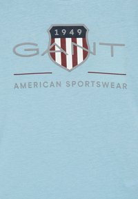 Lyseblå bomull T-skjorte med et skjoldlogo med røde og hvite striper og teksten "GANT 1949 AMERICAN SPORTSWEAR" under.