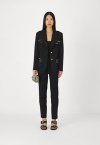 Costume noir avec boutons dorés, comprenant des poches avant. La veste ajustée est accompagnée de pantalons slim. Accessoirisé avec un petit sac vert et argent.