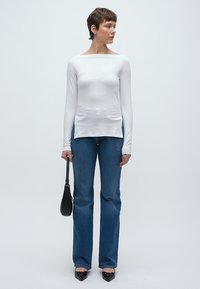 Fehér hosszú ujjú felső csónaknyakú kivitelben, kék bővülő szárú farmerral párosítva. A modell egy kis fekete kézitáskát tart, fekete lapos cipővel.