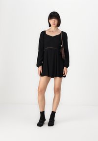 Melnā garās piedurknes kleita ar kvadrātveida dekoltē, pieguļošu augšdaļu ar mežģīņu apdari un platu svārku. Saskaņota ar melnām zeķēm un apaviem.