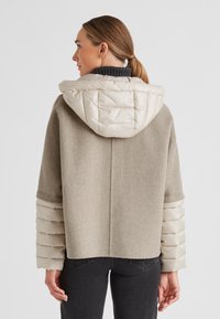 Beige Wollmischung Jacke mit einer puffy, hellbeigen Kapuze und Ärmeln. Hat einen kastigen Schnitt und einen gerippten Kragen sowie strukturierte Akzente an den Ärmeln.