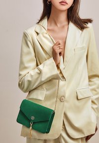 Borsa in suede verde con pattina pieghevole, tracolla in oro e dettaglio a nappina. Abbinata a un blazer in corduroy giallo chiaro.