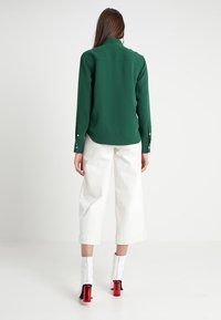 Blusa verde a maniche lunghe con colletto, abbinata a pantaloni bianchi larghi. Le calzature presentano alti tacchi rossi e zip.
