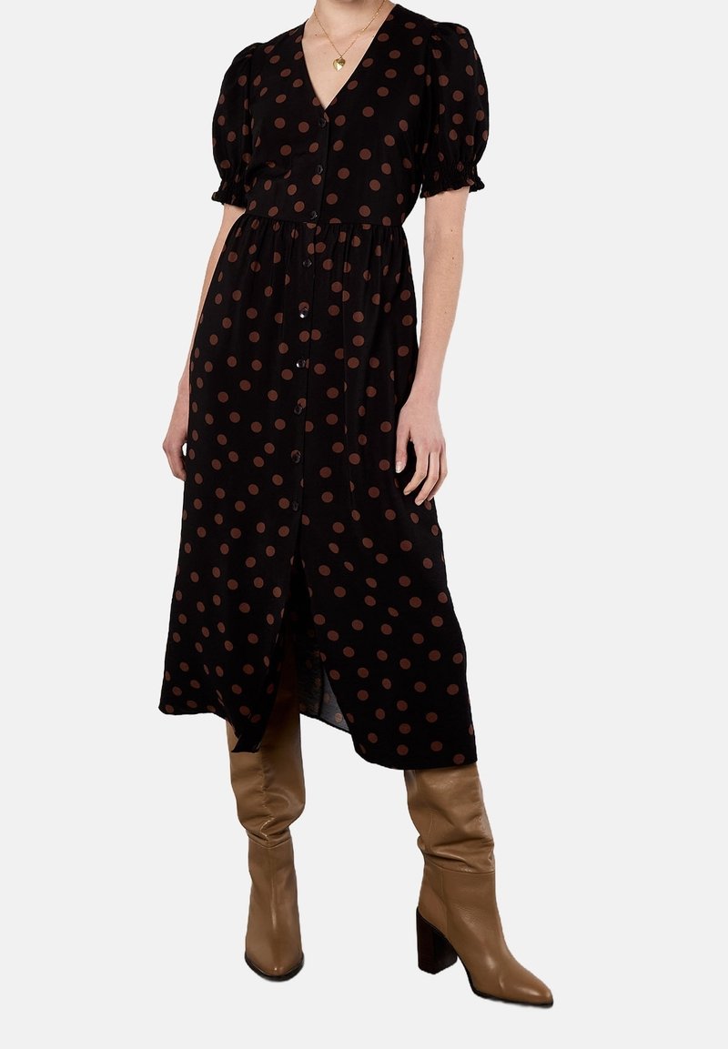 Femme portant une robe midi noire à pois marron, manches courtes bouffantes, boutons devant et bottes en cuir marron montant jusqu'aux genoux.