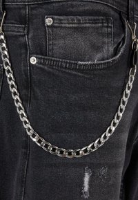 Los jeans de mezclilla negros presentan una cadena plateada sujeta al lazo del cinturón, con costuras visibles y pequeños detalles de desgaste en la tela.