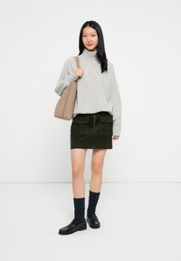 Siva pletena turtleneck pulover, zelena kordijska mini krila s žepi, mornarsko modre vzorčaste nogavice in črni loafer čevlji, z bež torbo.