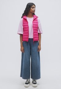 Ροζ καπιτονέ γιλέκο με κουκούλα πάνω από ανοιχτό ροζ μπλούζα, σετ με παντελόνι τζιν φαρδιάς γραμμής σε μπλε χρώμα και μαύρα-άσπρα αθλητικά παπούτσια. Ομοιόμορφα χρώματα σε όλο το σύνολο.