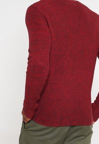 Punainen neulepaita, jossa on teksturoitu kuvio, pitkät hihat ja pyöreä kaula-aukko. Alaosa on hieman kaareva.