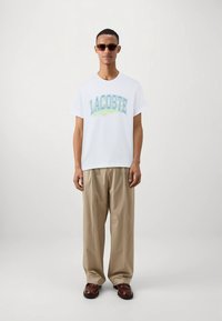 Balti medvilninė marškinėliai su turkio ir žalios spalvos "LACOSTE" grafika, derinama su smėlio spalvos kelnėmis ir rudeniniais rudais bateliais, modelis dėvi akinius nuo saulės.