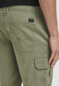 Pantalones cargo verde oliva de algodón con bolsillos de carga laterales, costuras visibles y una etiqueta de tela en el bolsillo trasero.