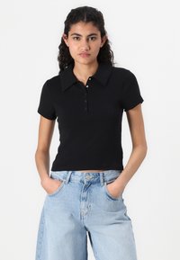 Svart polotröja med krage, korta ärmar och knäppning. Bärs tillsammans med ljusblå jeans, vilket visar en figurnära silhuett.