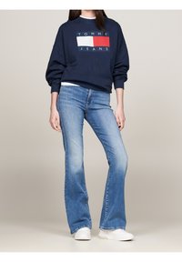 Žena v námořnickém svetru s obdélníkovým logem Tommy Jeans, světlých modrých zvonových džínách a bílých teniskách, stojící na neutrálním pozadí.