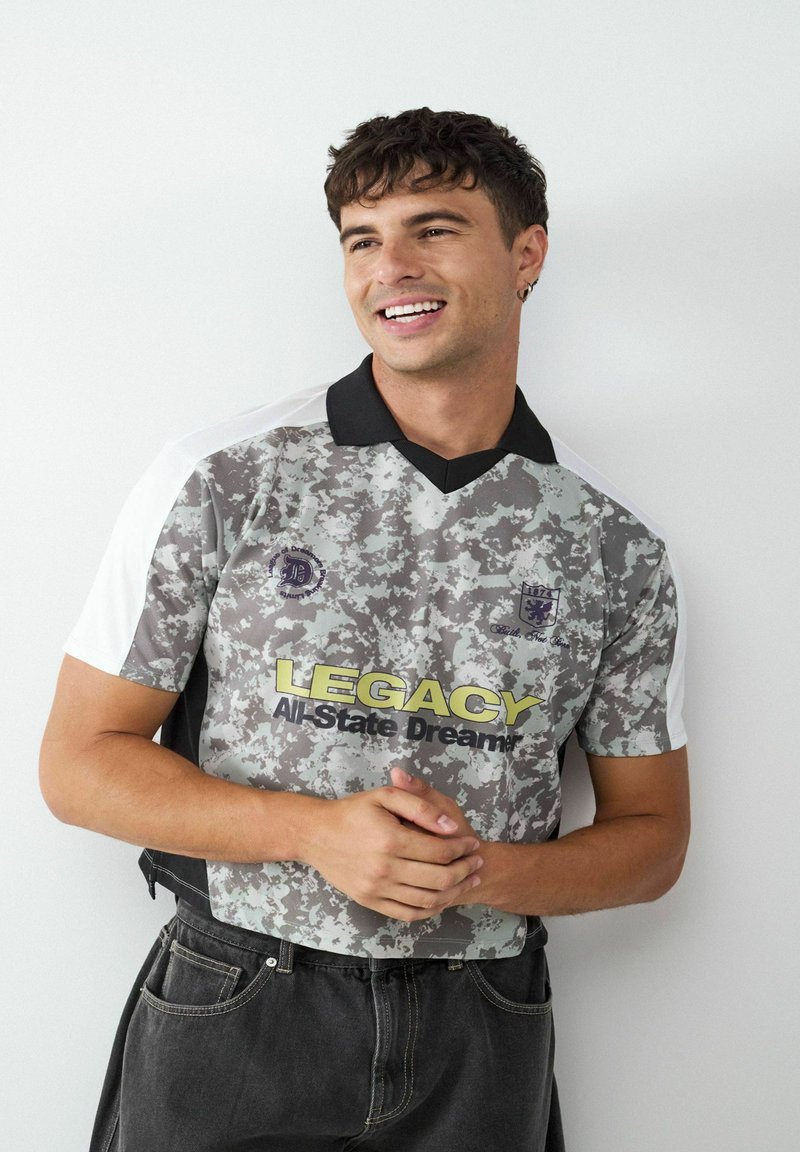 Szara koszulka z krótkim rękawem w wzór kamuflażu, z czarnym kołnierzykiem, z żółtym napisem "LEGACY" oraz haftowanymi logo na piersi.