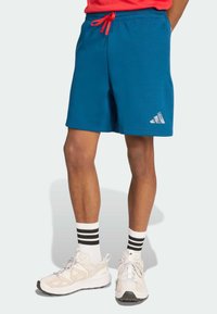 Blauwe sweatshirt-short met een rode trekkoord, voorzien van zijzakken en een wit logo op de zoom. Draagt bij witte schoenen en zwartgestreepte sokken.