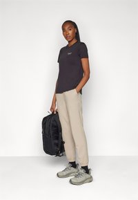 Schwarzes Baumwoll-T-Shirt mit kleinem Logo, beiger Jogginghose, grauen Wanderschuhen und einem schwarzen Rucksack mit strukturiertem Design.