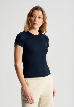 Kobieta o blond włosach do ramion, ubrana w dopasowaną granatową bluzkę z krótkim rękawem i jasnobeżowe spodnie, patrzy lekko w lewo.