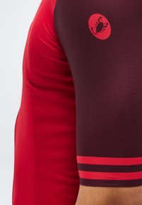 Κοντινό πλάνο ενός λυγισμένου βραχίονα που φοράει μαύρη και κόκκινη κοντομάνικη φανέλα ποδηλασίας με κόκκινο λογότυπο σκορπιού στο επάνω μανίκι.