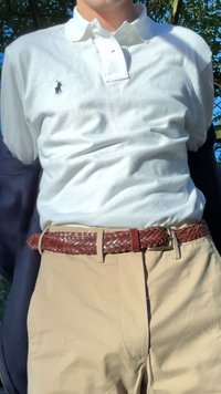 Tricou polo alb cu un mic logo bleumarin, asortat cu pantaloni bej și o curea împletită din piele maro. Croială confortabilă, materiale texturate vizibile.