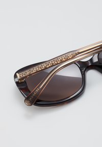 Gafas de sol de carey con lentes oscuros. La montura de acetato presenta un brazo con acentos en dorado y un patrón texturizado, así como el logo "COACH".