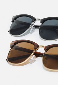  Dois pares de óculos de sol: um preto com detalhes prateados, o outro castanho com acabamentos dourados, apresentando almofadas nasais transparentes e formas arredondadas.