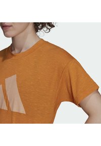Krátký rukáv oranžové tričko vyrobené z texturované tkaniny, s velkým béžovým logem na přední straně a kulatým výstřihem.
