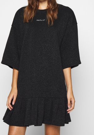 Kobieta ubrana w czarną, błyszczącą sukienkę z krótkim rękawem i falbanką na dole, z napisem "REPLAY" w kolorze białym na piersi.