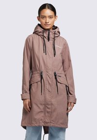 Femeie îmbrăcată cu o parka mauve cu glugă, cu sireturi, buzunare cu fermoar și blugi de culoare albastru deschis, stând în fața unui fundal luminos uniform.