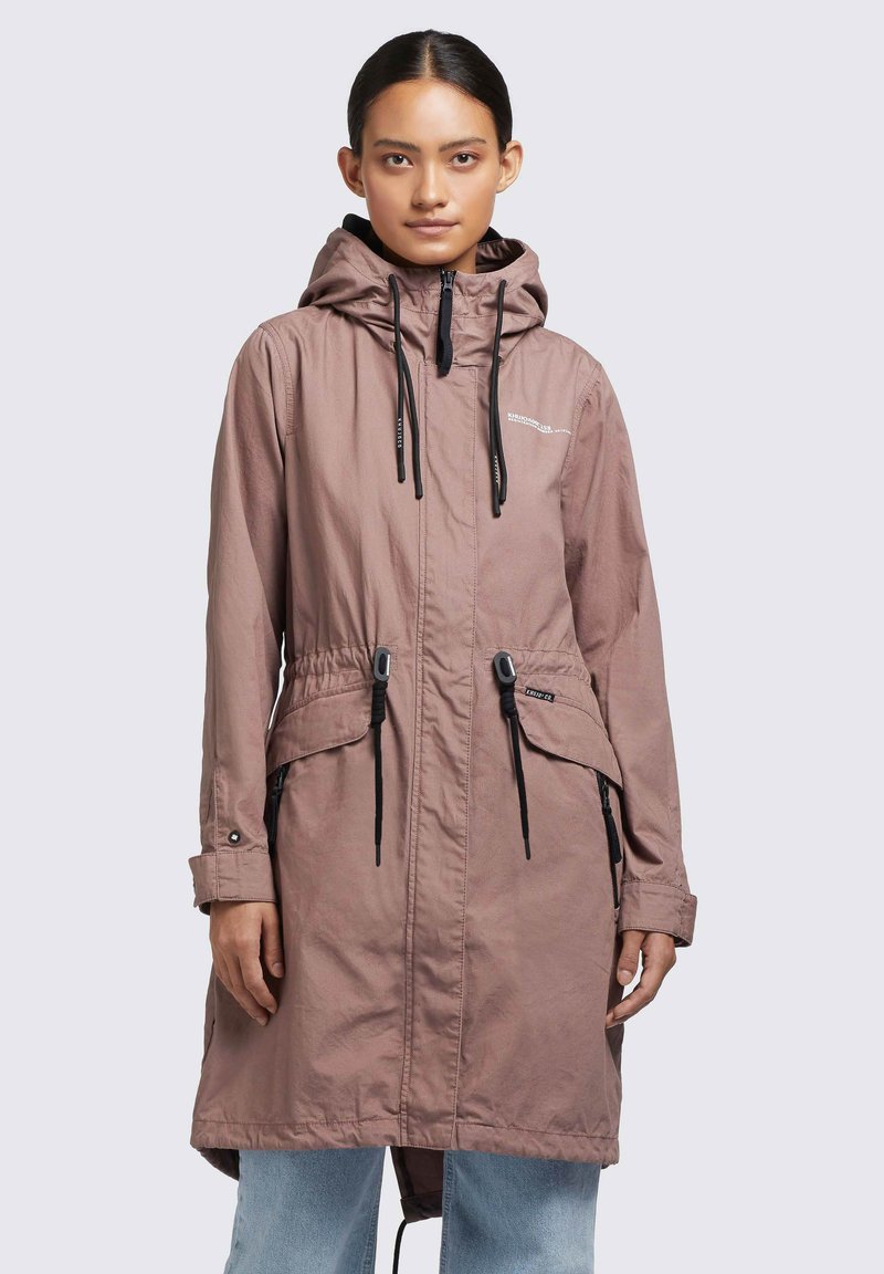 Femeie îmbrăcată cu o parka mauve cu glugă, cu sireturi, buzunare cu fermoar și blugi de culoare albastru deschis, stând în fața unui fundal luminos uniform.