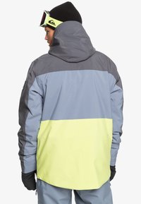 En skidjacka med en grå och gul färgblockdesign, med en huva, texturerad yta och elastiska ärmmuddar för en säker passform.