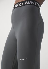Šedé sportovní legíny s černým pasem označeným "NIKE PRO." Mají hladkou texturu a bílý Nike swoosh logo na dolní části nohy.
