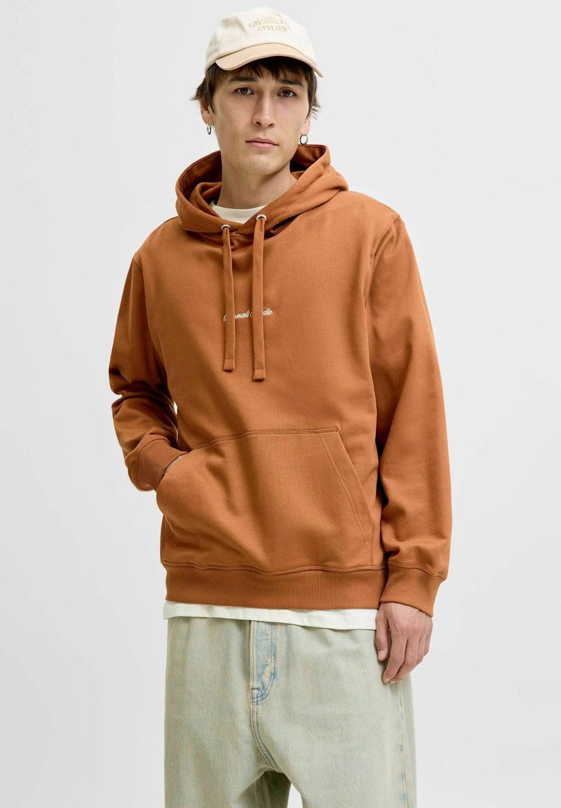 Brun hoodie med en framficka, snoddar och vit broderad text. Kombinerad med ljusa denimbyxor och en beige keps.