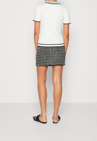 Witte top met korte mouwen en een zwarte bies, gecombineerd met een zwart-witte tweed minirok. Model draagt zwarte sandaaltjes zonder bandje achter de hiel.