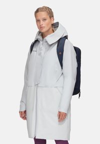 Veste longue imperméable gris clair avec capuche, présentant une fermeture éclair à l'avant, des poches latérales et un design superposé. Portée sur un sac à dos noir.