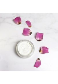 Vit kräm i en genomskinlig burk med ett swirl-mönster, omgiven av rosa rosenblad på en vit bakgrund.