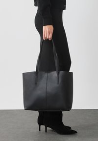 Sac fourre-tout en cuir noir avec deux longues poignées, texture lisse et détail de couture verticale. Tenue par une personne portant des vêtements noirs et des bottes à talons.