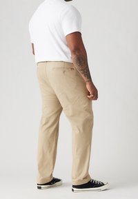 Beżowe bawełniane spodnie o prostym kroju nogawki, z detalem tylnych kieszeni i subtelną metką marki. Noszone z czarnymi trampkami dla kontrastu.