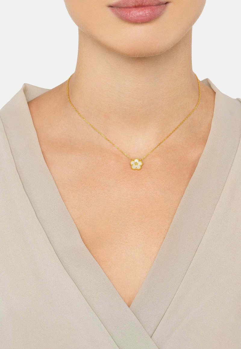 Gold necklace featuring a floral pendant with a white accent, worn on a neck with a light beige, v-neck shirt.