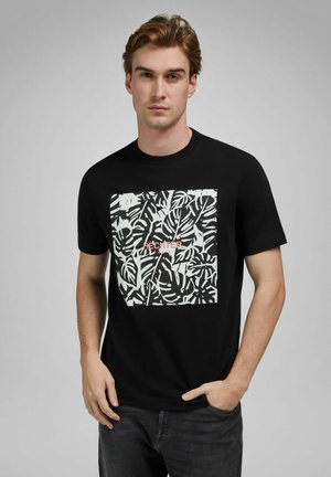 Junger Mann mit hellbraunen Haaren steht frontal, trägt ein schwarzes T-Shirt mit weißem tropischem Blattmuster und dem Schriftzug "Hechter Paris".